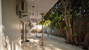 A covered walkway with a sandy floor, trees, and a brick wall. - Velana Vibes – Your Tranquil Getaway in the Heart of Mathiveri, Maldives (Mathiveri)