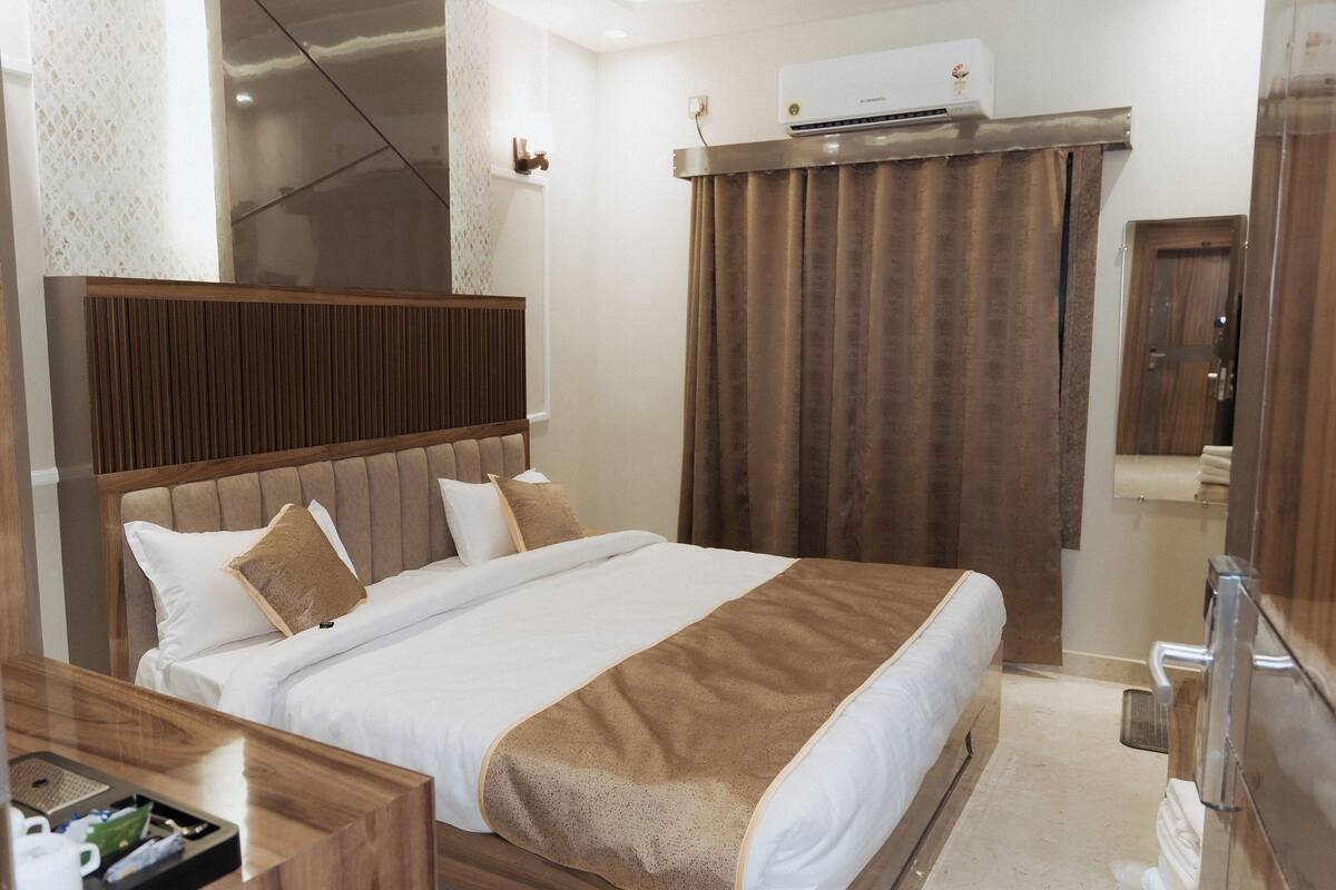 Une chambre d’hôtel avec un lit, une tête de lit en bois, un plaid marron, une table de chevet avec une lampe et un appareil de climatisation.