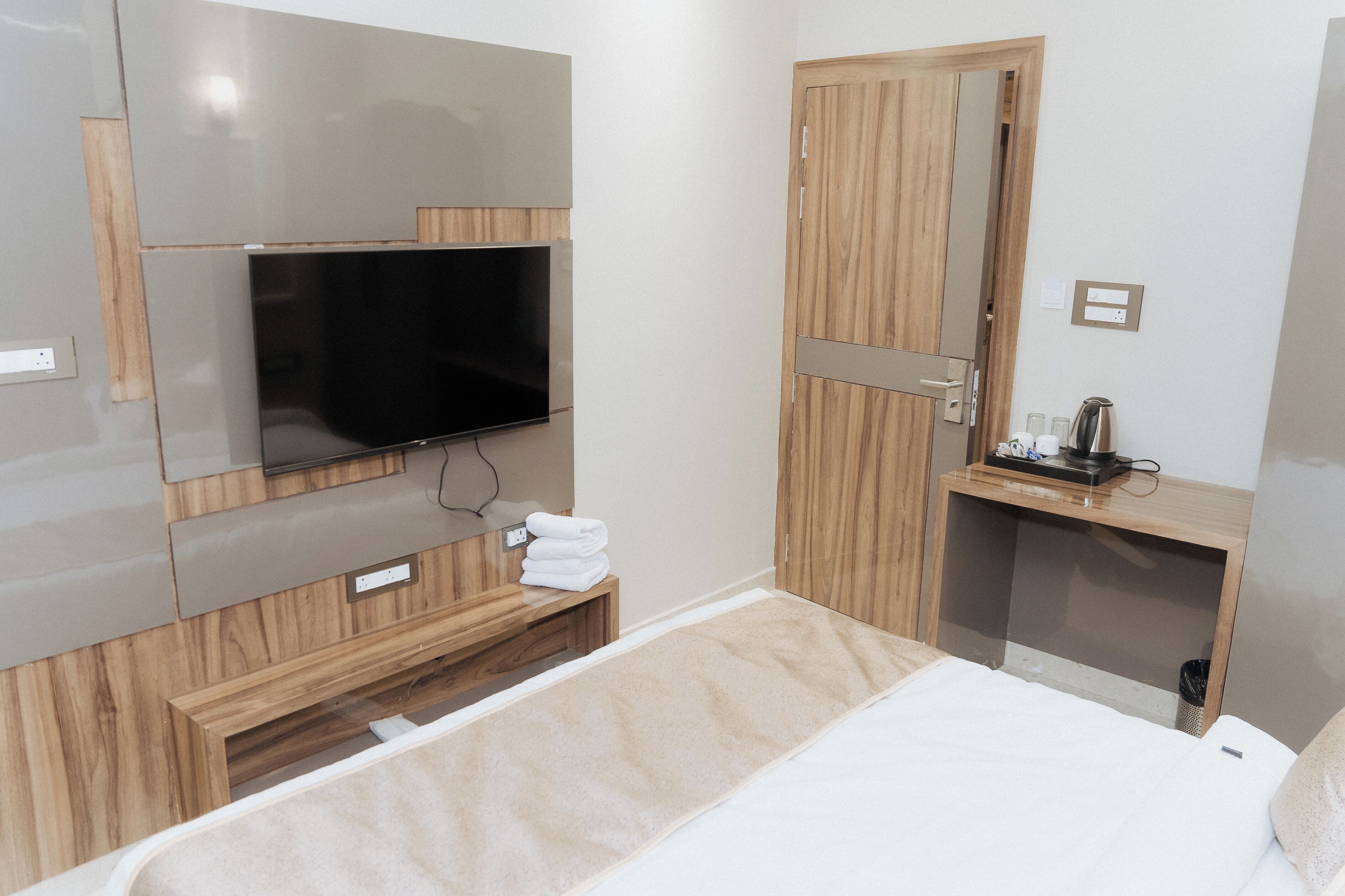 Una camera d'albergo moderna con TV a schermo piatto montata a parete, un armadio in legno, un comodino con bollitore e un letto rifatto con cura.