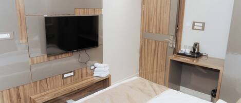 Quarto de hotel moderno com televisão de tela plana na parede, guarda-roupa de madeira, mesa de cabeceira com chaleira e cama bem arrumada.
