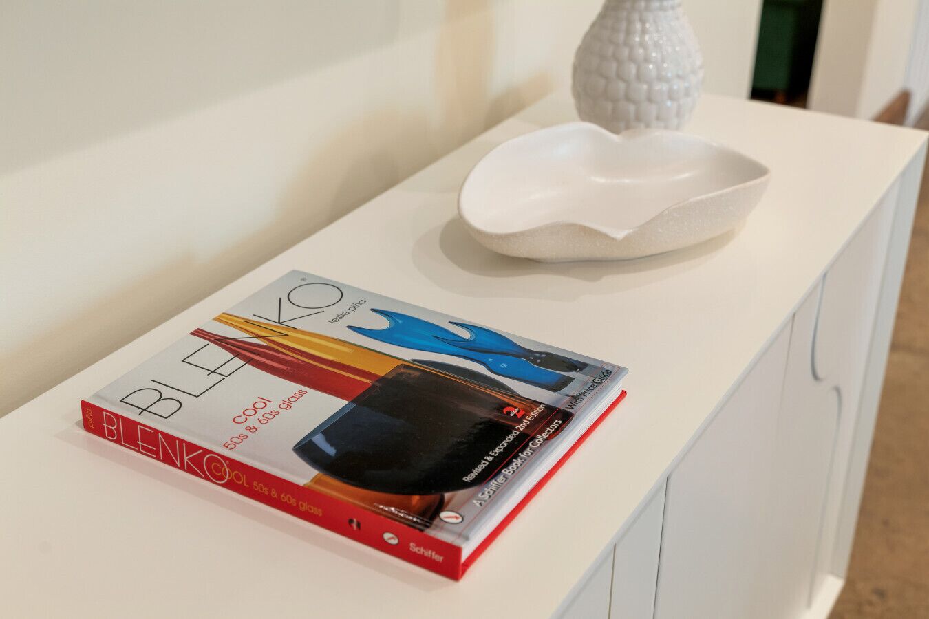 Una mesa blanca con un libro titulado "BLENKO COOL 50s & 60s gloss" y un cuenco decorativo blanco encima.