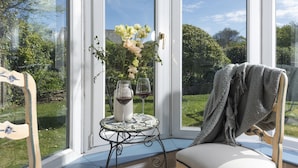 Een kamer met een groot raam, een tafeltje met een vaas en wijnglazen, en een stoel met een plaid.