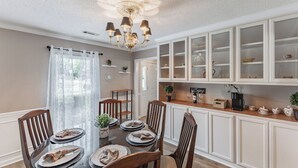 Un comedor con una mesa redonda dispuesta para cuatro personas, rodeada de sillas de madera. Hay una araña sobre la mesa, y una zona de cocina con armarios blancos y una cafetera en la encimera.
