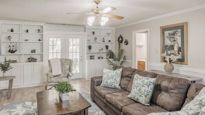 Una sala de estar con un sofá de cuero marrón, un sillón blanco y una mesa de centro con una planta. Hay estanterías con objetos decorativos y un ventilador de techo.