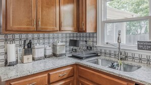 Una cocina con armarios de madera, encimera de granito, ventana con vistas y un letrero que dice 'Mom's Kitchen Abierto las 24 Horas'.