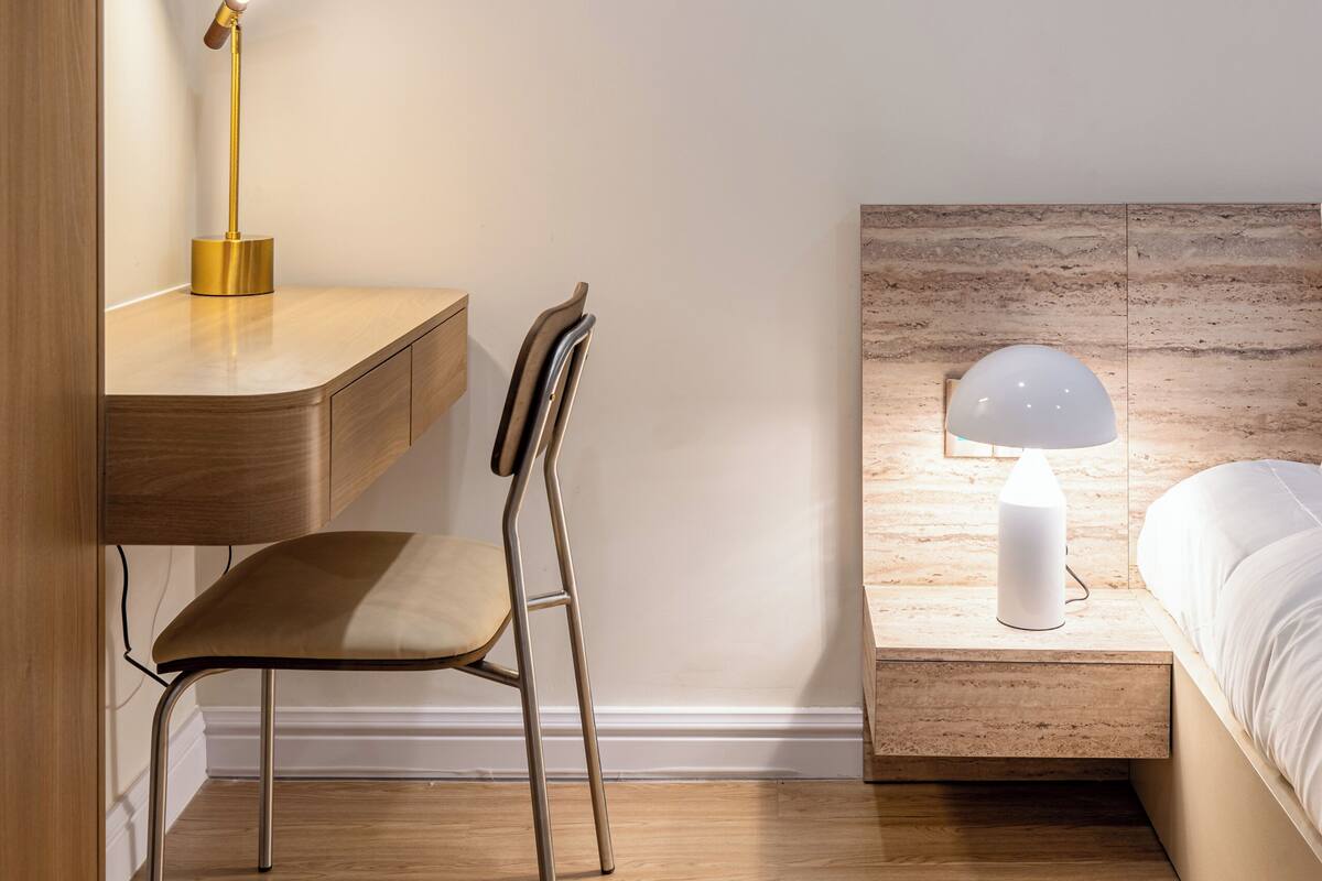 A bedroom with a wooden desk, a chair, a bedside table with a lamp, and a bed.