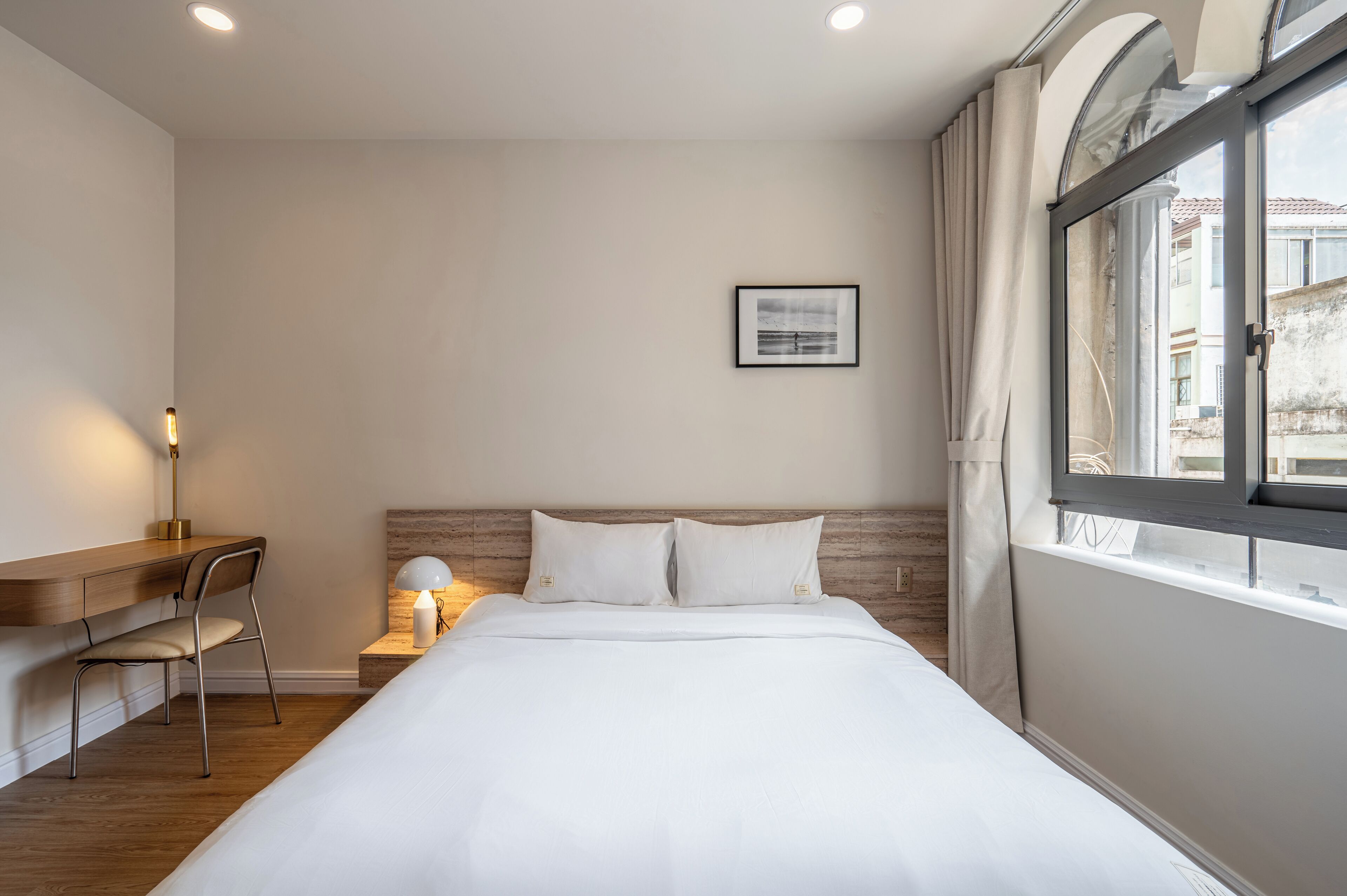 Une chambre à coucher comprenant un lit, un bureau, une chaise, une table de chevet avec une lampe et une fenêtre avec des rideaux.