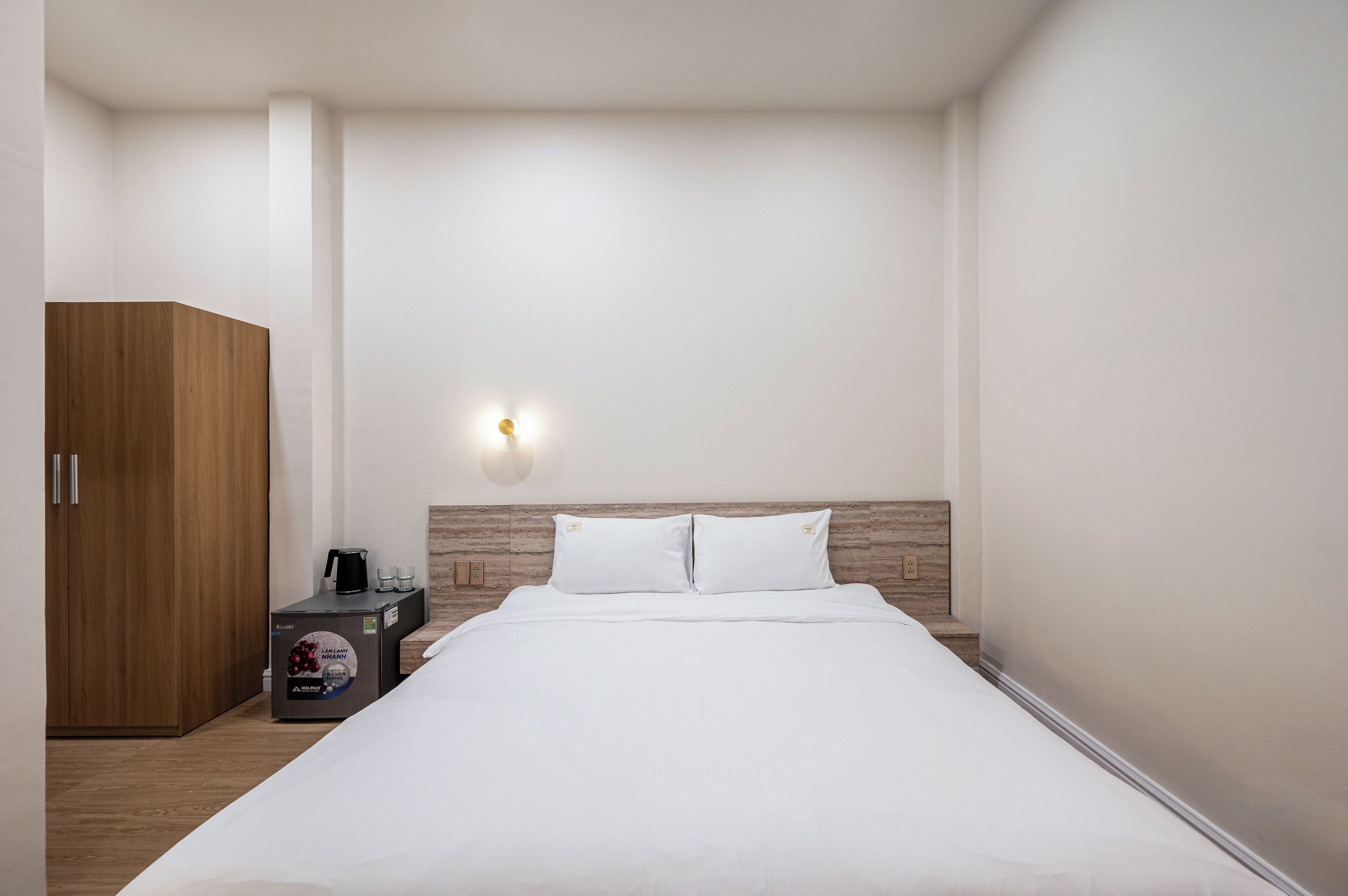 A modern hotel room with a large bed, a wooden wardrobe, a coffee machine, and a wall-mounted light fixture.