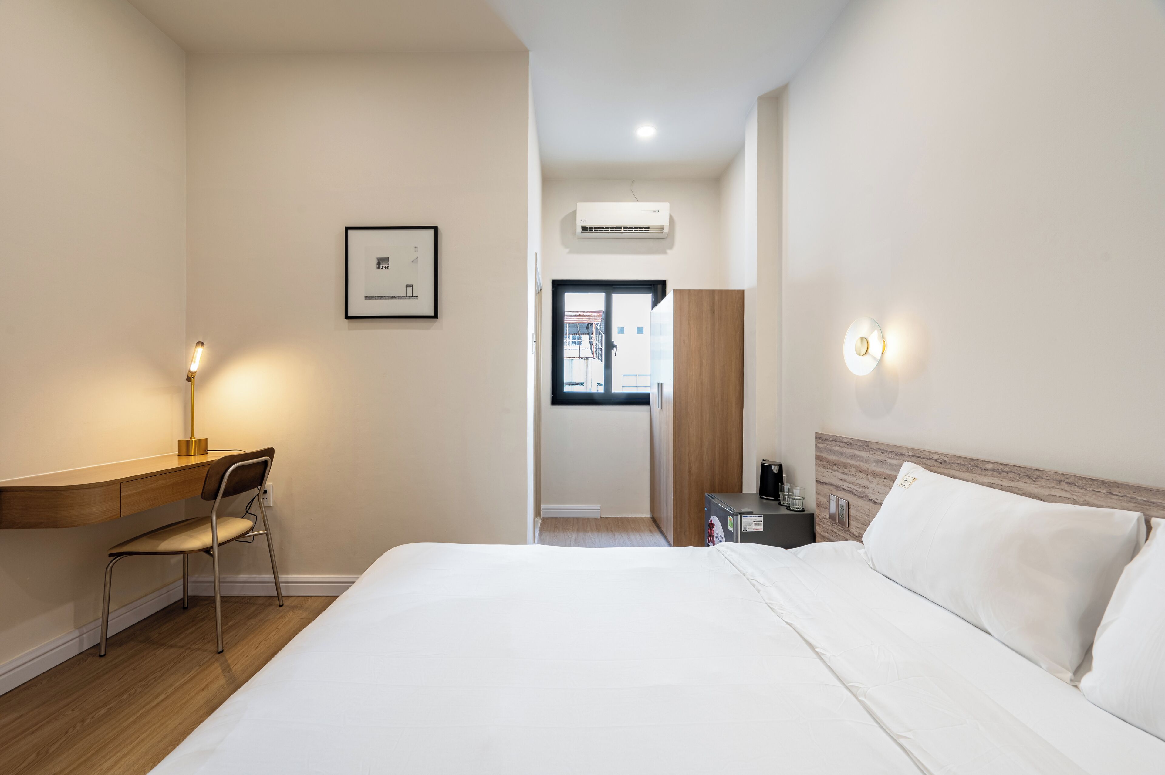 A hotel room with a bed, desk, chair, bedside table, and an air conditioning unit.