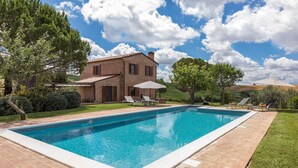 Una piscina con una casa de ladrillo al fondo, rodeada de vegetación y un cielo despejado.