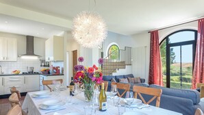 Una cocina moderna con armarios blancos, un comedor con mesa dispuesta para seis personas, y un ventanal con vistas al campo.