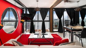 A modern living room with a red accent wall, a round window, a dining area with a table set for two, and a city view.
