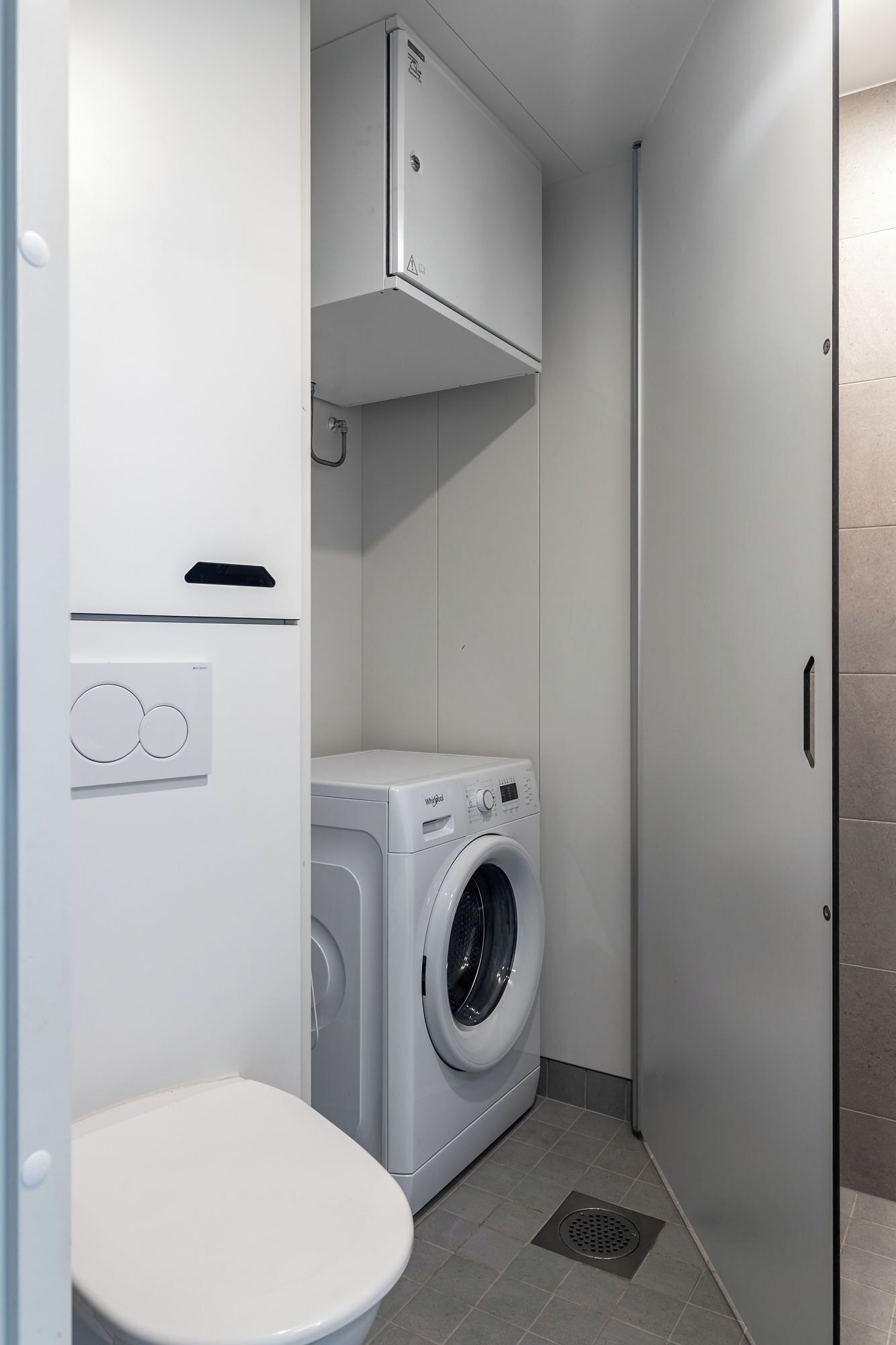A compact bathroom with a white toilet, a washing machine, and a cabinet above the machine.