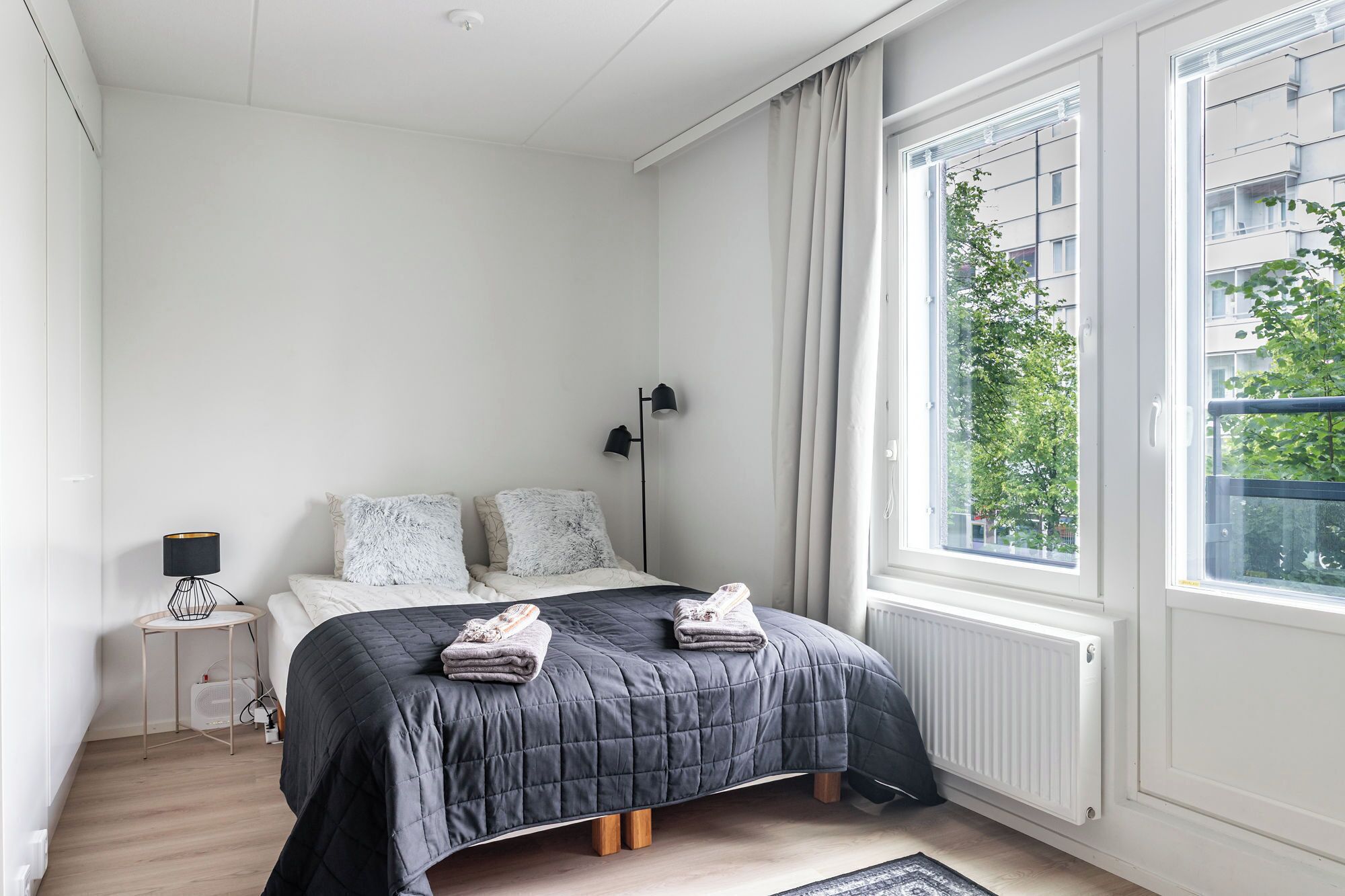 Une chambre à coucher avec un lit, une table de chevet, une lampe et une fenêtre donnant sur des immeubles et des arbres.