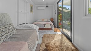 A compact bedroom with two beds, a wardrobe, a small table, and a large window offering a view of the outdoors.
