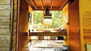 A wooden cabin interior with a wooden ceiling, a sink, a mirror, and a window overlooking greenery.
