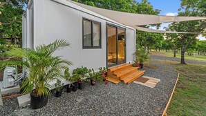 Una casa móvil compacta y moderna con entrada por puerta corrediza de vidrio, un pequeño porche con escalones de madera y diversas plantas en macetas alrededor del exterior.