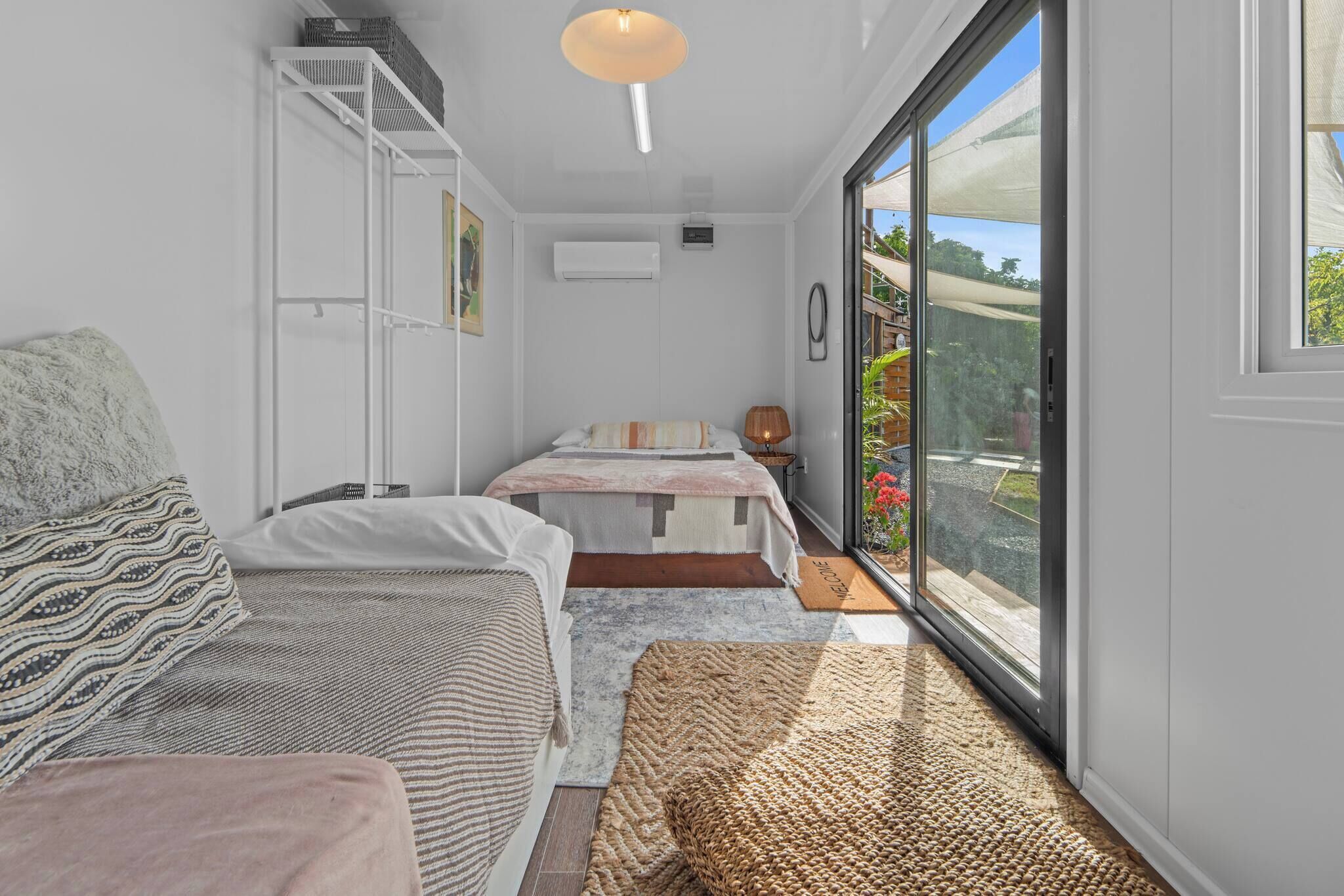 A compact bedroom with two beds, a wardrobe, a small table, and a large window offering a view of the outdoors.