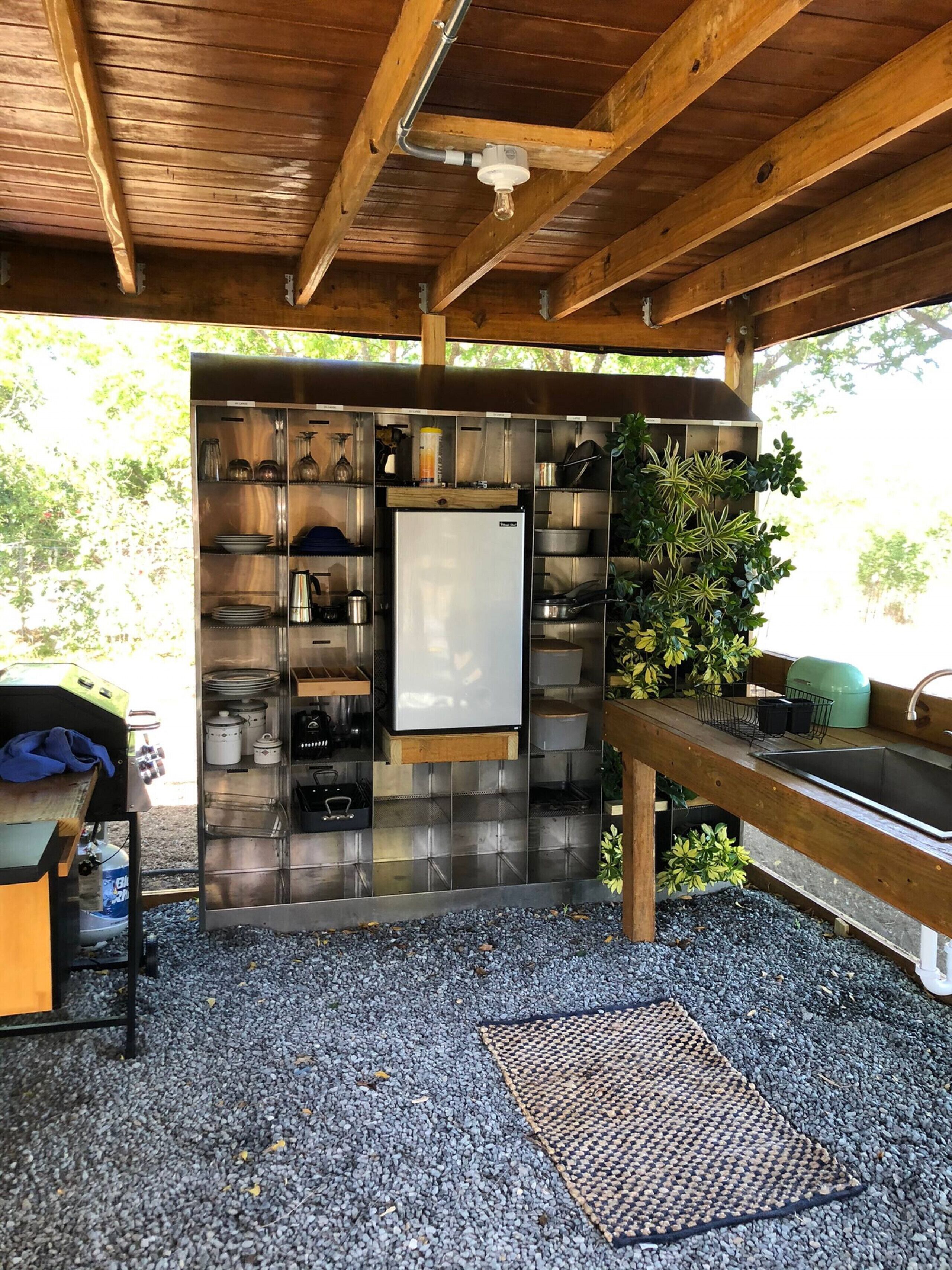 A compact kitchen with a built-in refrigerator, stainless steel shelves, and a sink under a wooden roof.