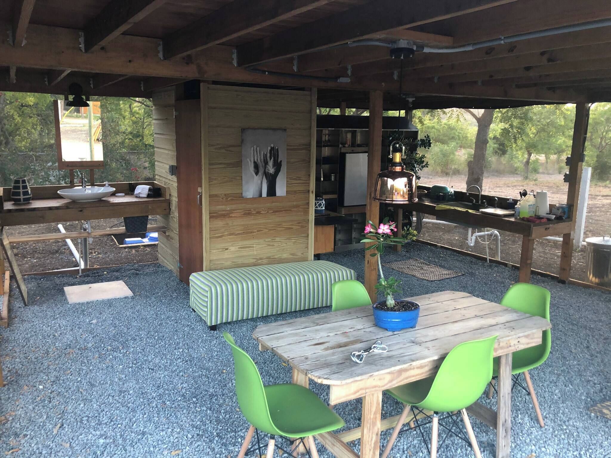 Un patio al aire libre con una mesa de madera, sillas verdes, un banco, un lavamanos y una parrilla.