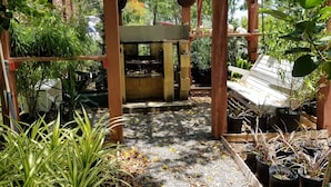 Eine Holz-Pergola mit Grillplatz aus Backsteinen darunter, umgeben von Topfpflanzen und einem Kiesweg.