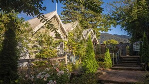 A traditional building with a steeply pitched roof, surrounded by lush greenery and a stone bridge.