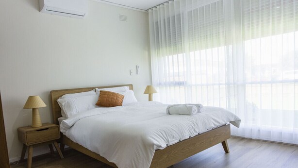 A neatly made bed with white linens, a wooden headboard, and a single brown pillow. There are two bedside tables with lamps, one on each side of the bed. The room has a large window with sheer curtains, allowing natural light to enter.