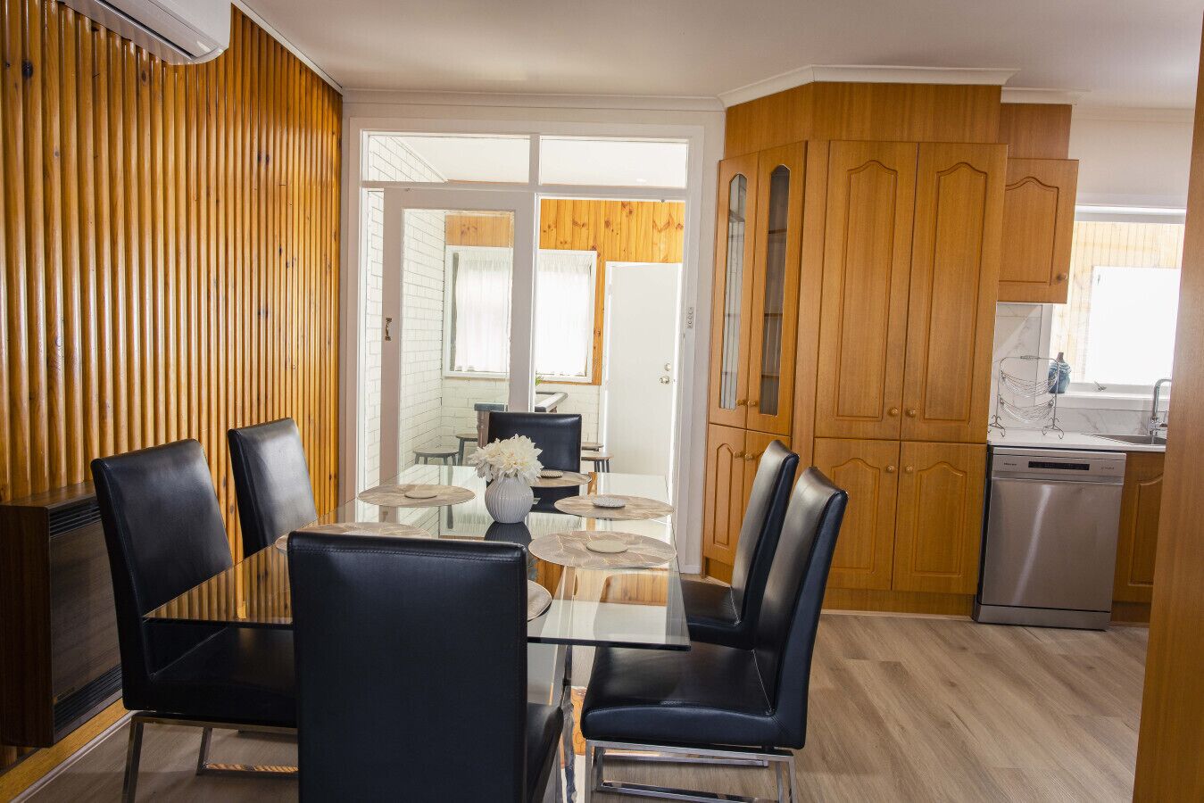 Une cuisine moderne avec des meubles en bois, un lave-vaisselle et un coin repas comprenant une table en verre et des chaises noires.