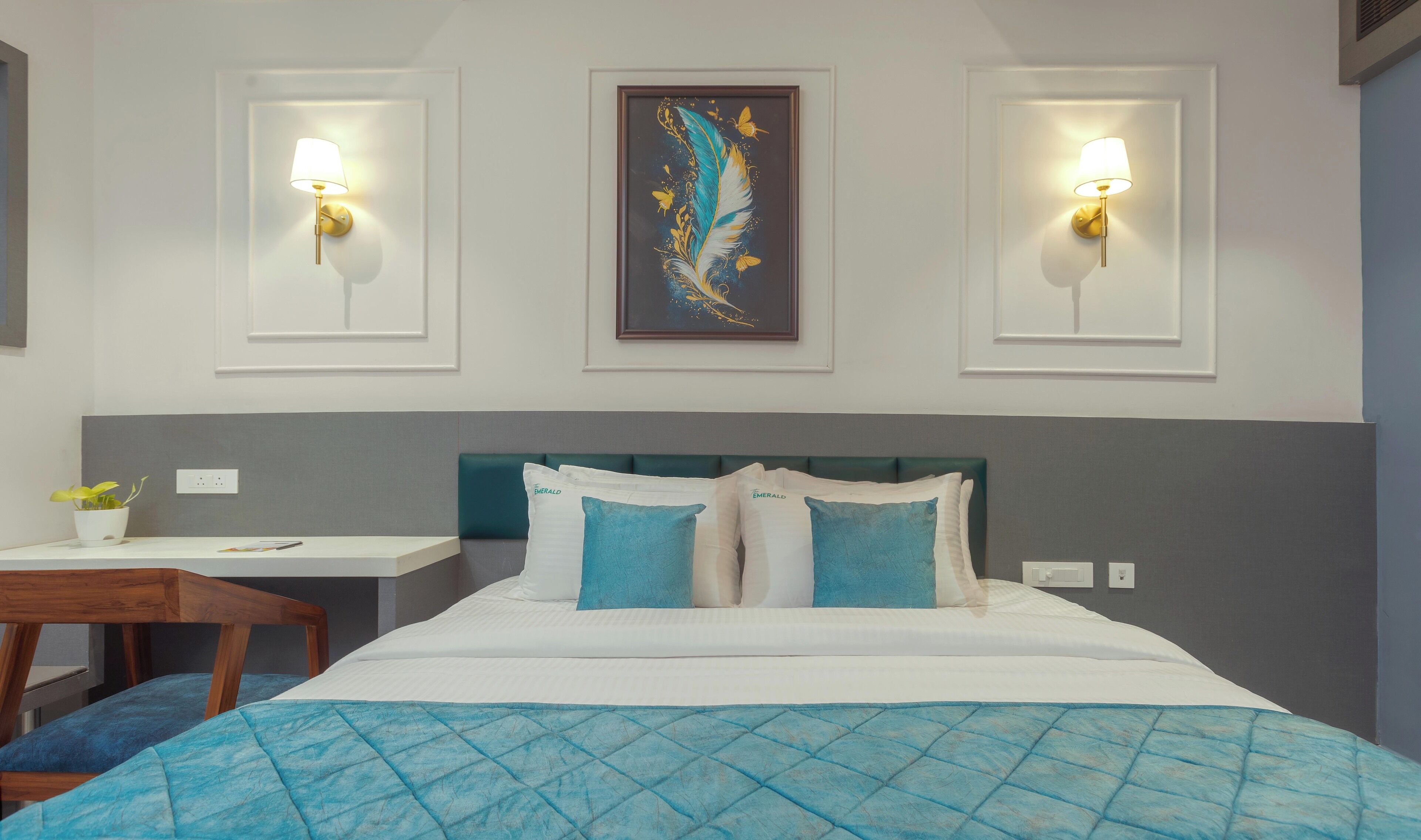 A hotel room with a bed, two wall-mounted lamps, a framed artwork, a small table with a plant, and a blue quilted bedspread.