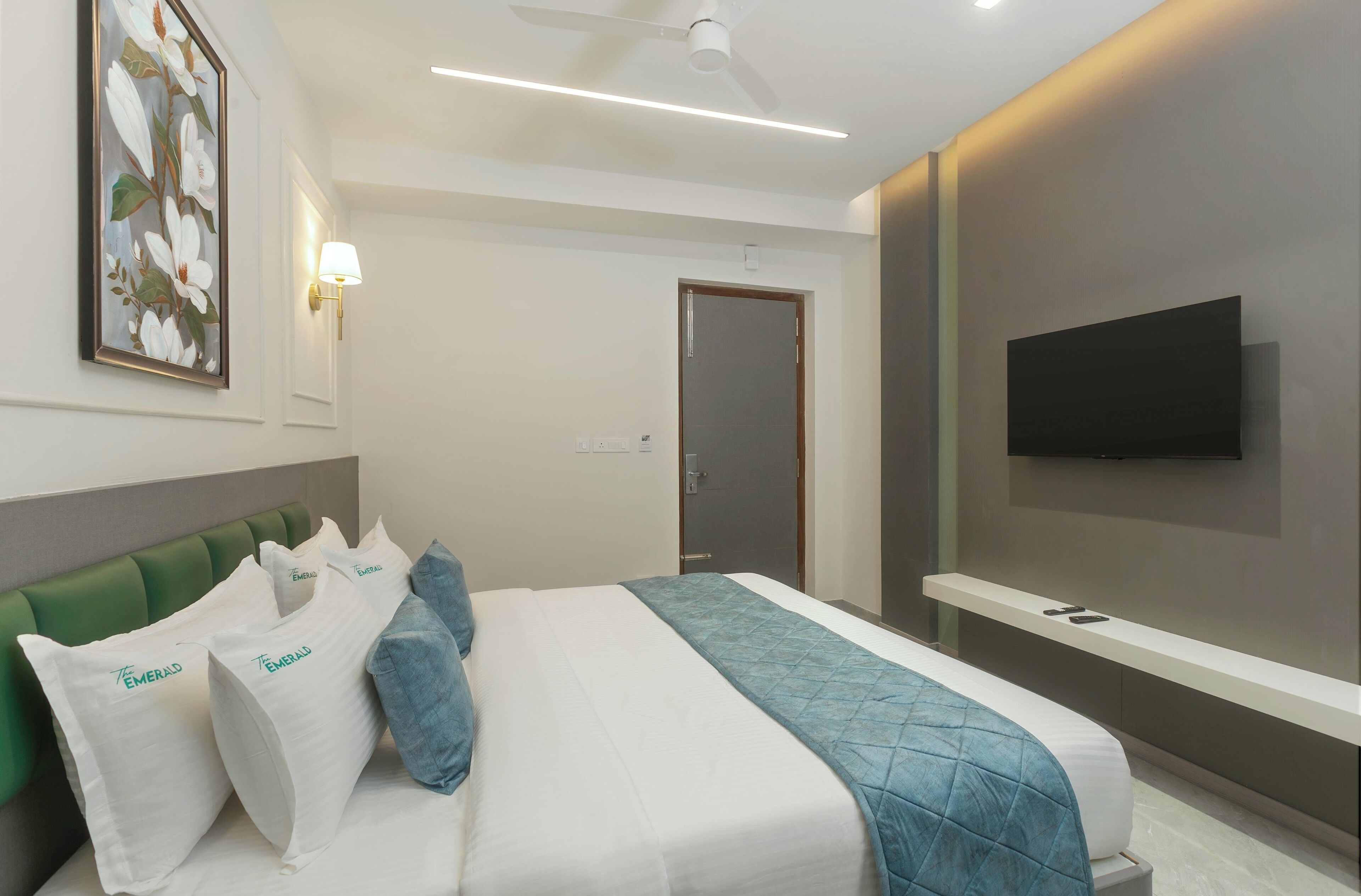 Una habitación de hotel moderna con cama, cabecero tapizado en verde, ropa de cama blanca, una manta acolchada azul y un cuadro con motivos florales en la pared.