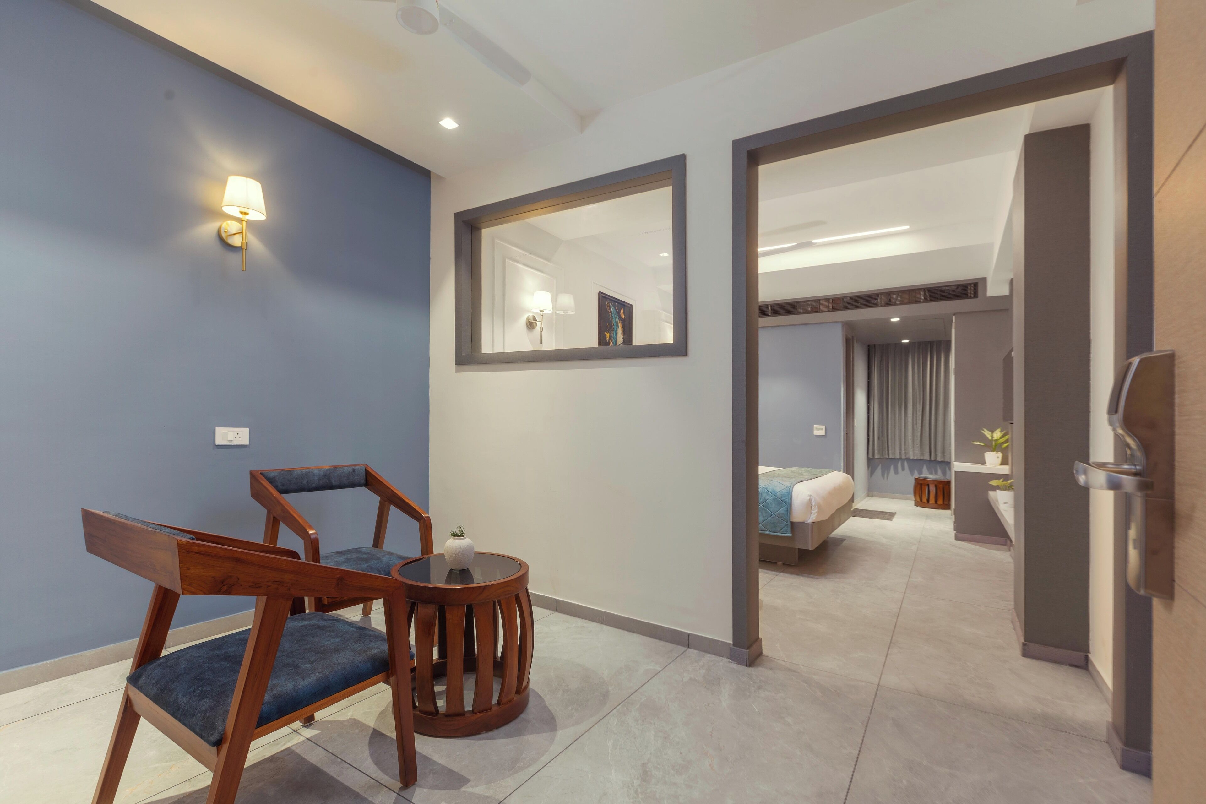 Una habitación de hotel moderna con una pared azul decorativa, una silla de madera, una mesa redonda, un espejo grande y una cama con colcha azul.