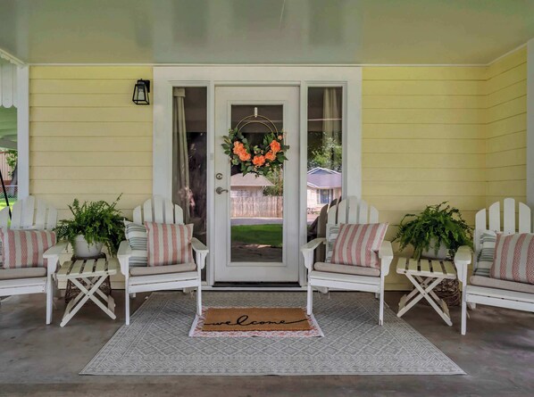 A porch with two white Adirondack chairs, a welcome mat, and a potted plant.