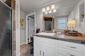 A bathroom with a large mirror, a sink, and a vanity with drawers. There is a towel rack and a shower visible in the background.