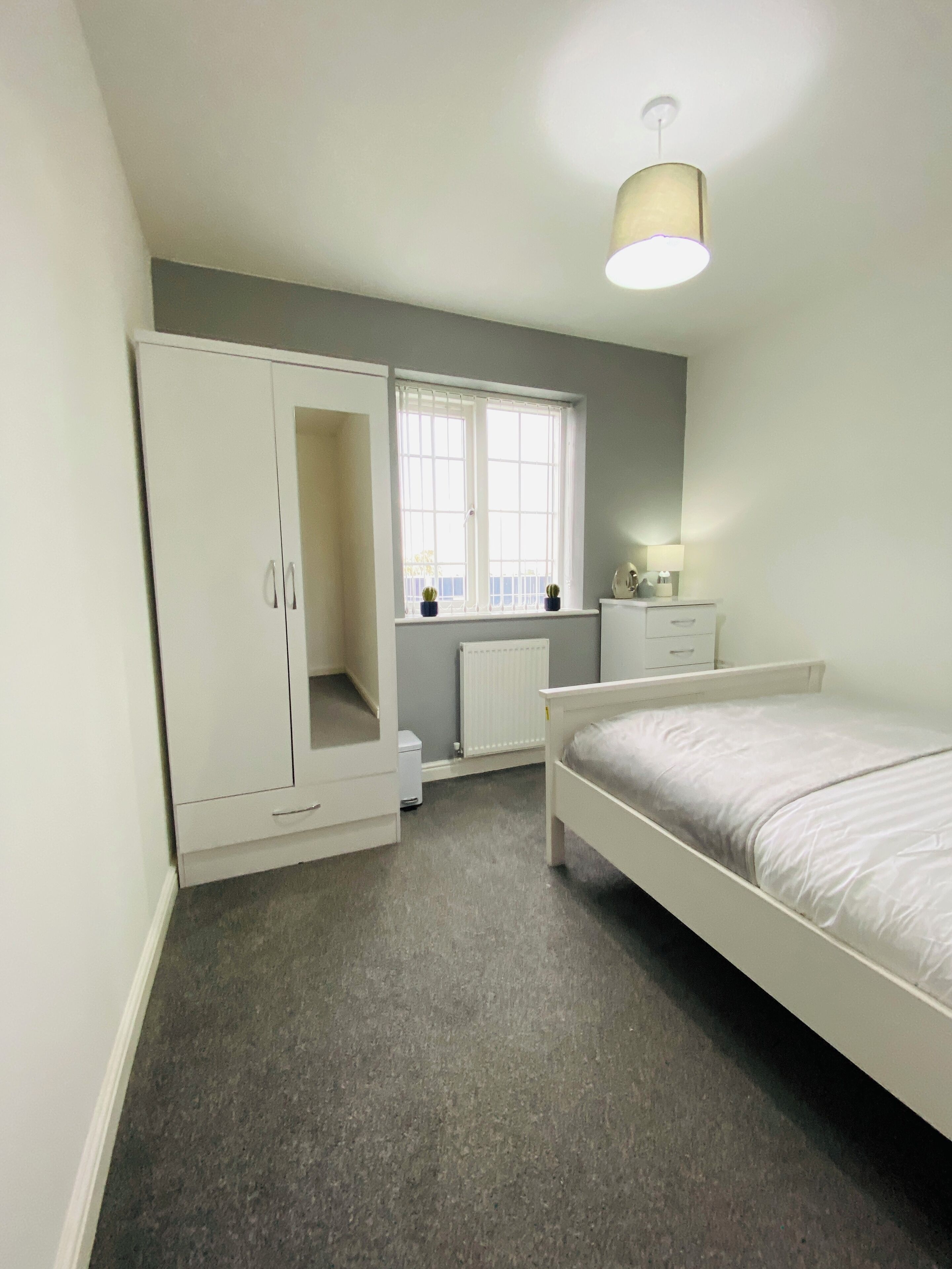 A bedroom with a bed, wardrobe, radiator, window, and a lamp.