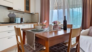 Un coin repas avec une table dressée pour deux personnes, une cuisine en arrière-plan et une vue à travers des rideaux légers.