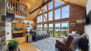 Un vaste espace de vie avec une grande fenêtre donnant sur un lac, un coin salon confortable avec un canapé et des fauteuils, et une cuisine agrémentée d’éléments en bois.