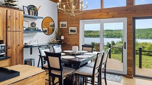 Un intérieur en bois comprenant une table à manger, des chaises, un lustre et une vue sur un lac, visible à travers de grandes fenêtres.