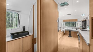 Interior moderno de una caravana con cocina, que incluye un lavabo negro, armarios de madera y una ventana con vistas a árboles.