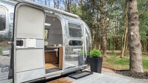 Una caravana plateada estacionada en una zona boscosa con una terraza y una pequeña planta en maceta.