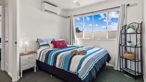 Un dormitorio con cama, mesita de noche, ventana con cortinas, ventilador de techo y estante metálico negro con una planta.