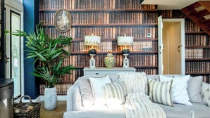 A cozy living room with a large bookshelf filled with books, a comfortable sofa, and a potted plant. - Campden Barn - Luxury Home with Hot Tub (Chipping Campden)