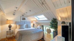 A bedroom with a large bed, a skylight, a potted plant, and a bedside table with a lamp. - Campden Barn - Luxury Home with Hot Tub (Chipping Campden)