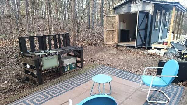 Un espace extérieur rustique aménagé avec un coin salon, un barbecue, deux chaises bleues et un tapis à motifs.