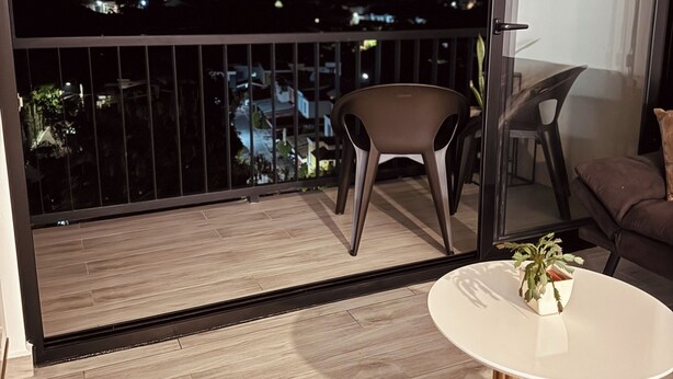 A balcony with a view of a cityscape at night, a round table with a plant, and two chairs. - Modern Condo - Beautiful City & Volcano Views - 5 min to WTC & Top Places (San Salvador)