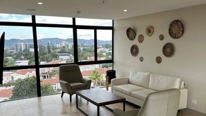 A spacious living room with a large window offering a city view, a white sofa, a coffee table, and decorative wall art. - Modern Condo - Beautiful City & Volcano Views - 5 min to WTC & Top Places (San Salvador)