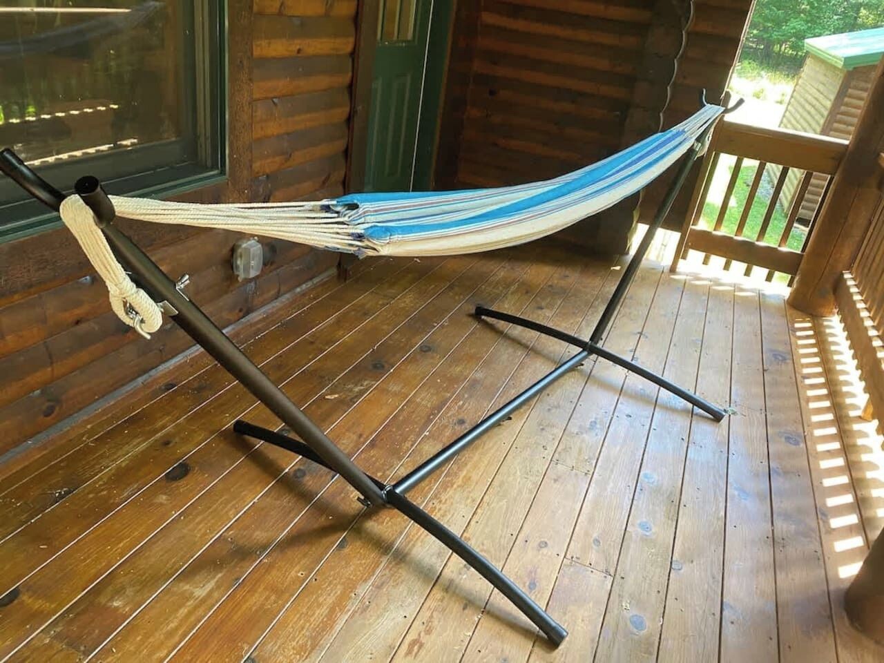 Un hamac sur une terrasse en bois, avec des supports métalliques, une fenêtre et une porte en arrière-plan.