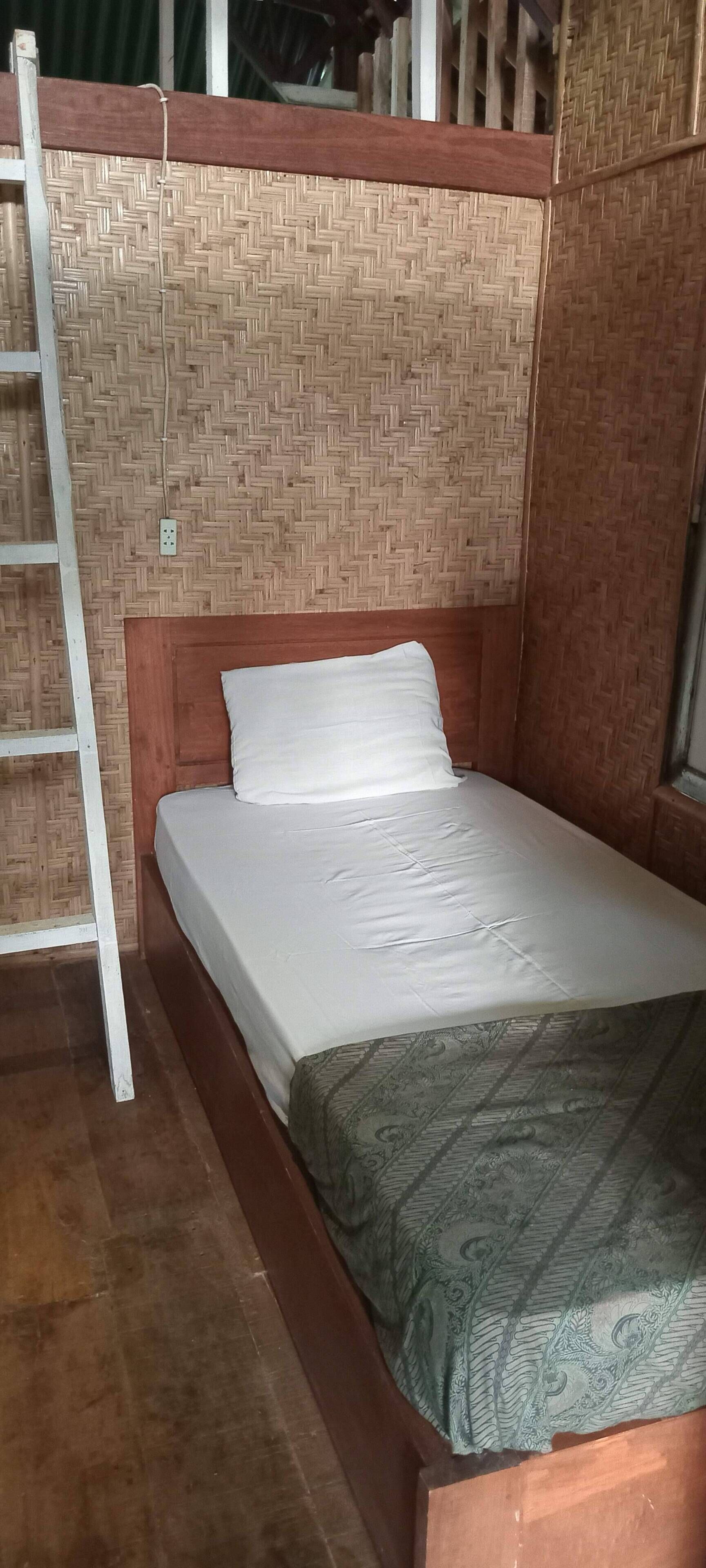 Un lit simple avec une tête de lit en bois, un oreiller blanc et une couverture à motifs. Une échelle est appuyée contre le mur, et un panneau mural tissé est visible.
