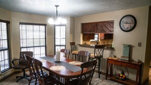Uma sala de jantar com mesa de madeira, cadeiras e um lustre. Há janelas com persianas, uma área de cozinha com armários e um relógio na parede.