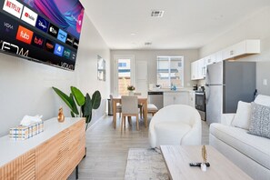 Ein modernes Wohnzimmer mit Flachbildfernseher, der verschiedene Streaming-Dienste anzeigt, einem Essbereich mit Tisch und Stühlen, einer Küche mit weißen Schränken und einem Kühlschrank sowie einem großen Fenster, das für natürliches Licht sorgt.