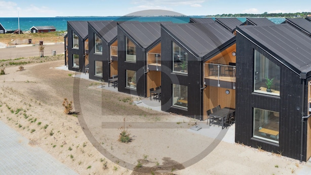 A row of modern, dark-colored cabins with large windows and wooden decks, situated by a beach with a clear view of the sea and a distant hill. - Havnehusene, Lejl. 59 (Slagelse)
